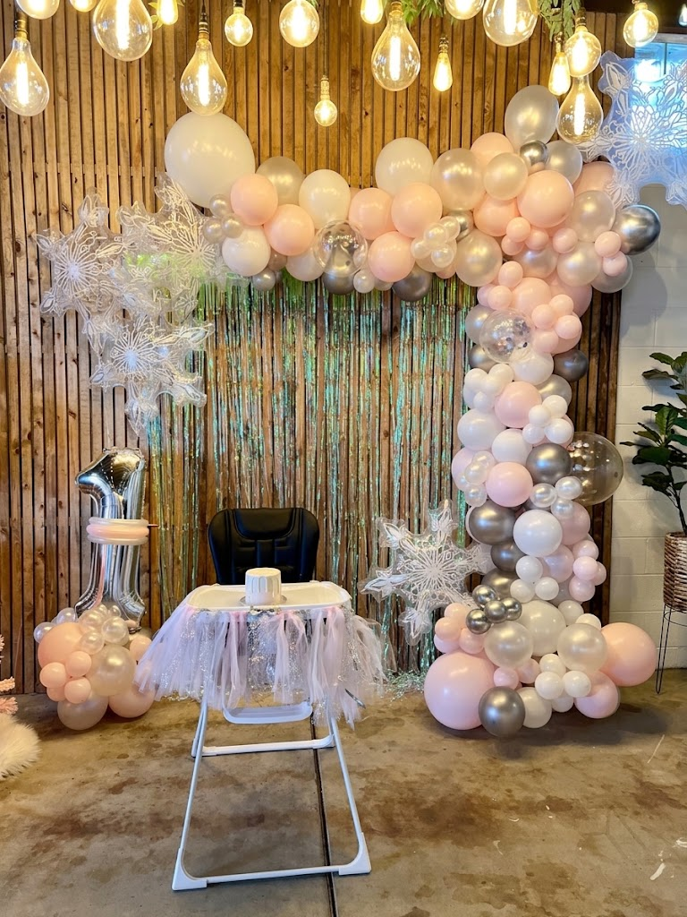 An organic balloon arch in blush, white and silver with snowflake accents over an iridescent tinsel backdrop beside a high chair first-birthday setup