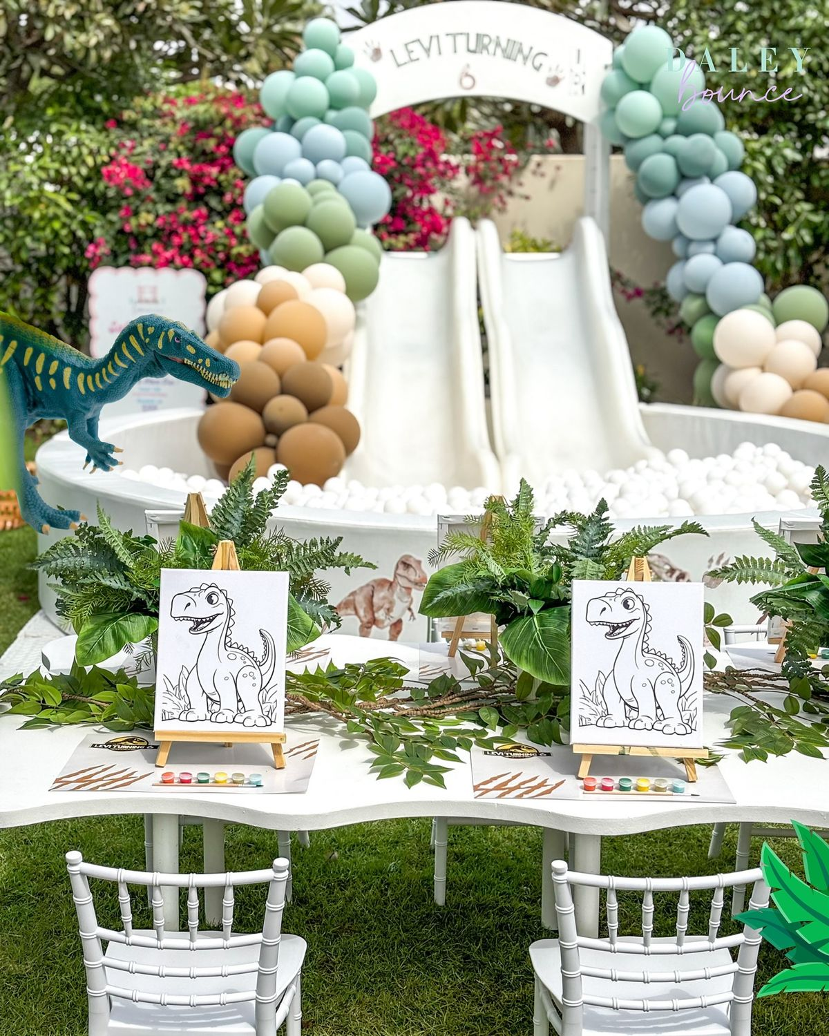 Dinosaur garden celebration with a white ball pit and twin slides behind a leafy kids’ activity table