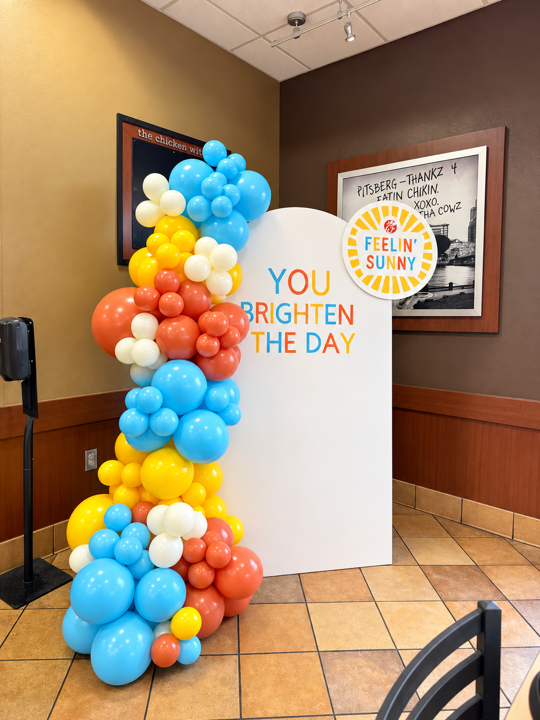 Blue, yellow, cream and terracotta balloon garland on a white arch backdrop reading You Brighten the Day