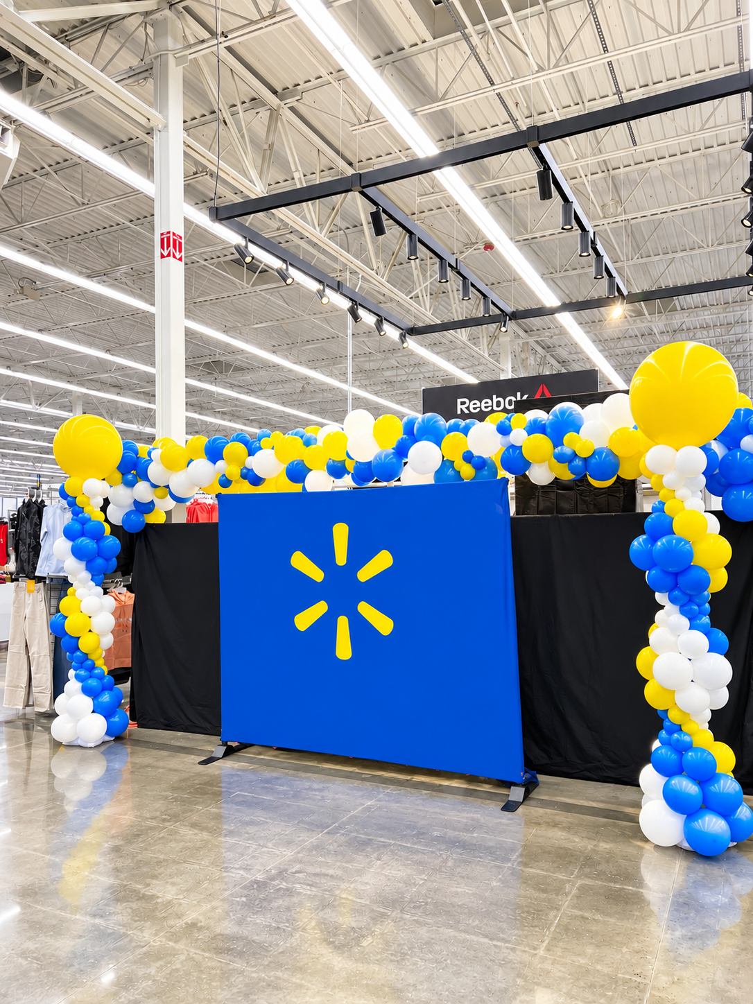 A blue branded backdrop with a yellow spark logo flanked by blue, yellow and white balloon columns at a corporate event