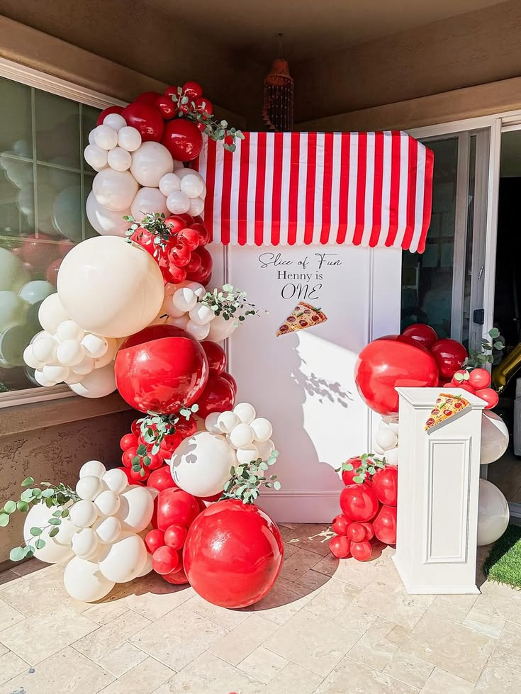 Red and white balloon arch around a pizza-party backdrop with striped awning and eucalyptus